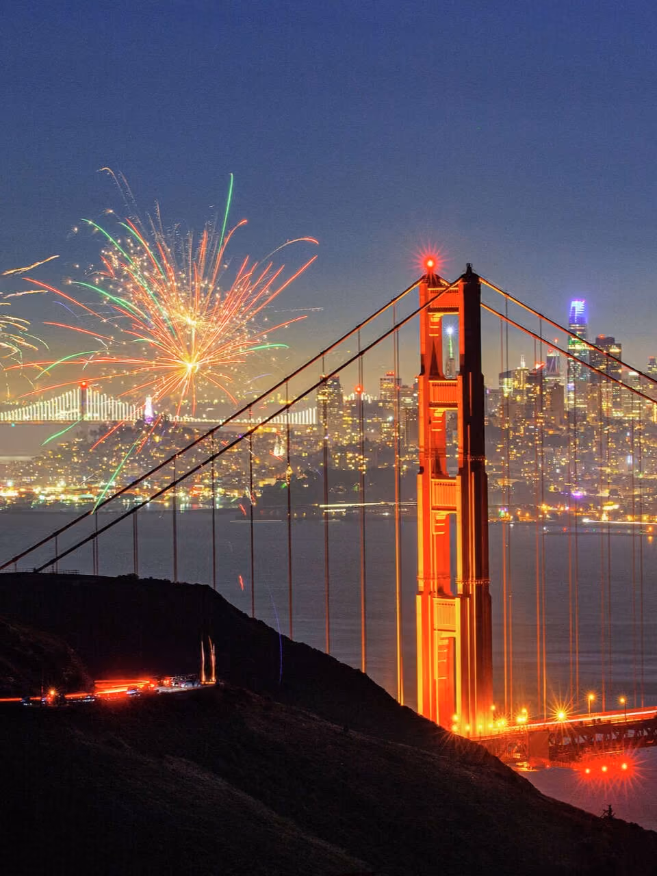 Golden Gate Bridge at night with fireworks bursting over San Francisco city skyline.