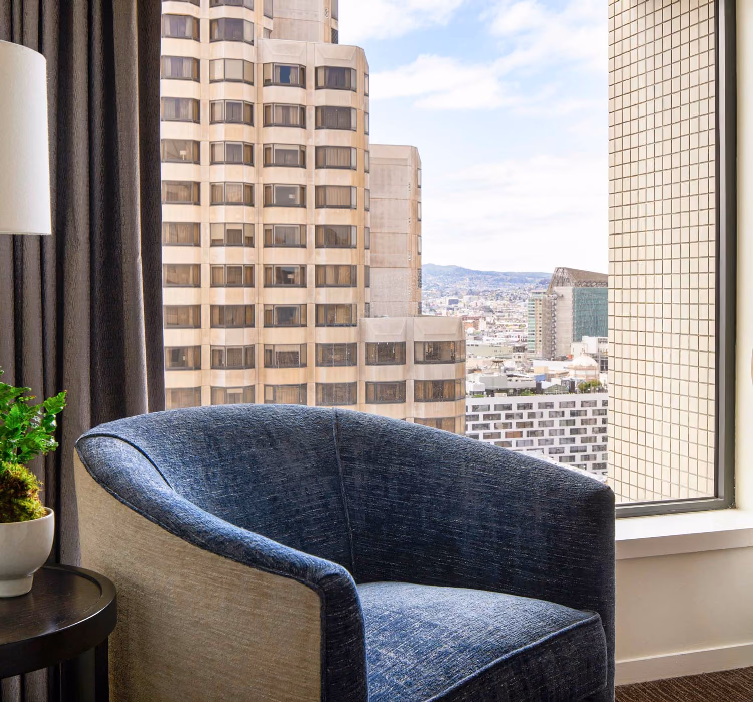 A dark blue cushioned chair by a window with a city view of tall buildings and hills.