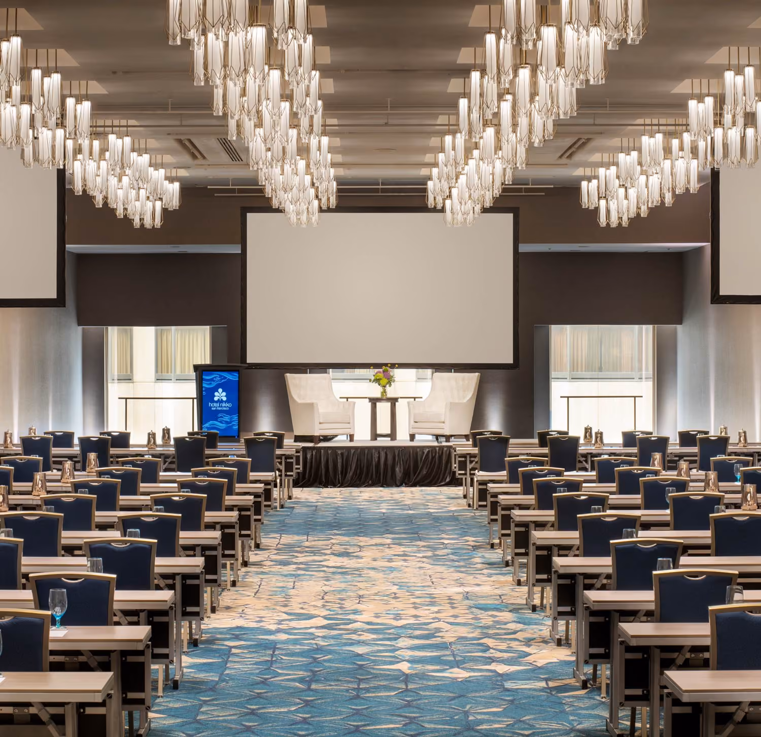Spacious conference room with rows of tables and chairs facing a stage with two white armchairs and a large screen.