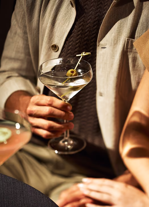 Person holding a martini glass with an olive garnish, sitting next to another person.