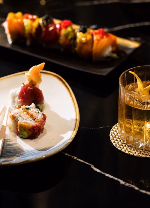 Two plates of assorted sushi rolls and a glass of whiskey with lemon twist on a dark marble table.