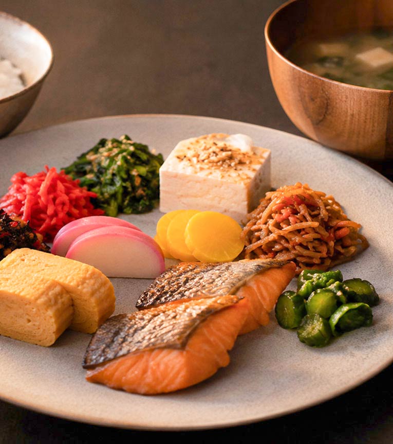 Japanese breakfast plate with grilled salmon, tamagoyaki, tofu, pickles, and vegetables on a light plate.