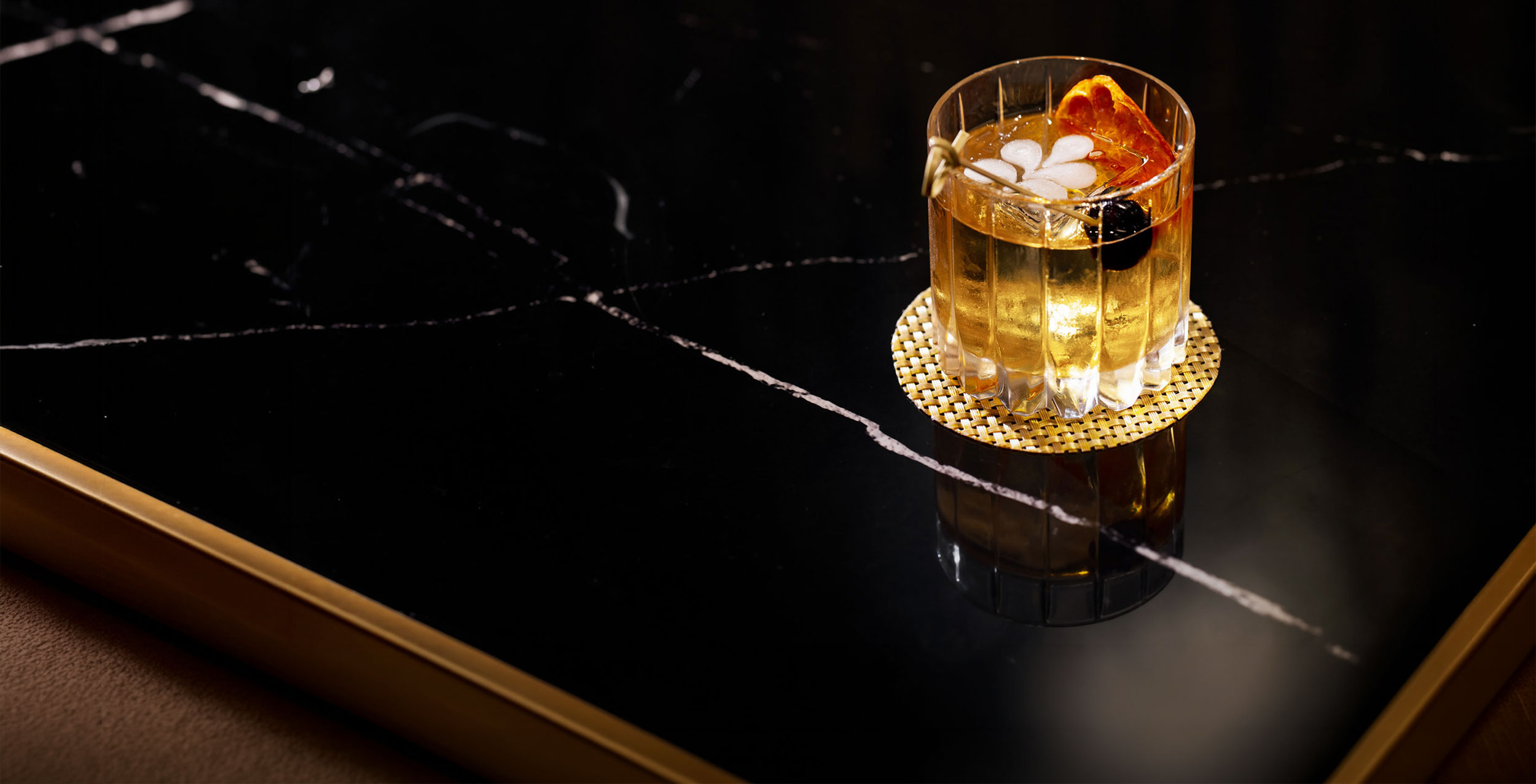 Glass of amber cocktail with ice, an orange slice, and a cherry on a black marble table.