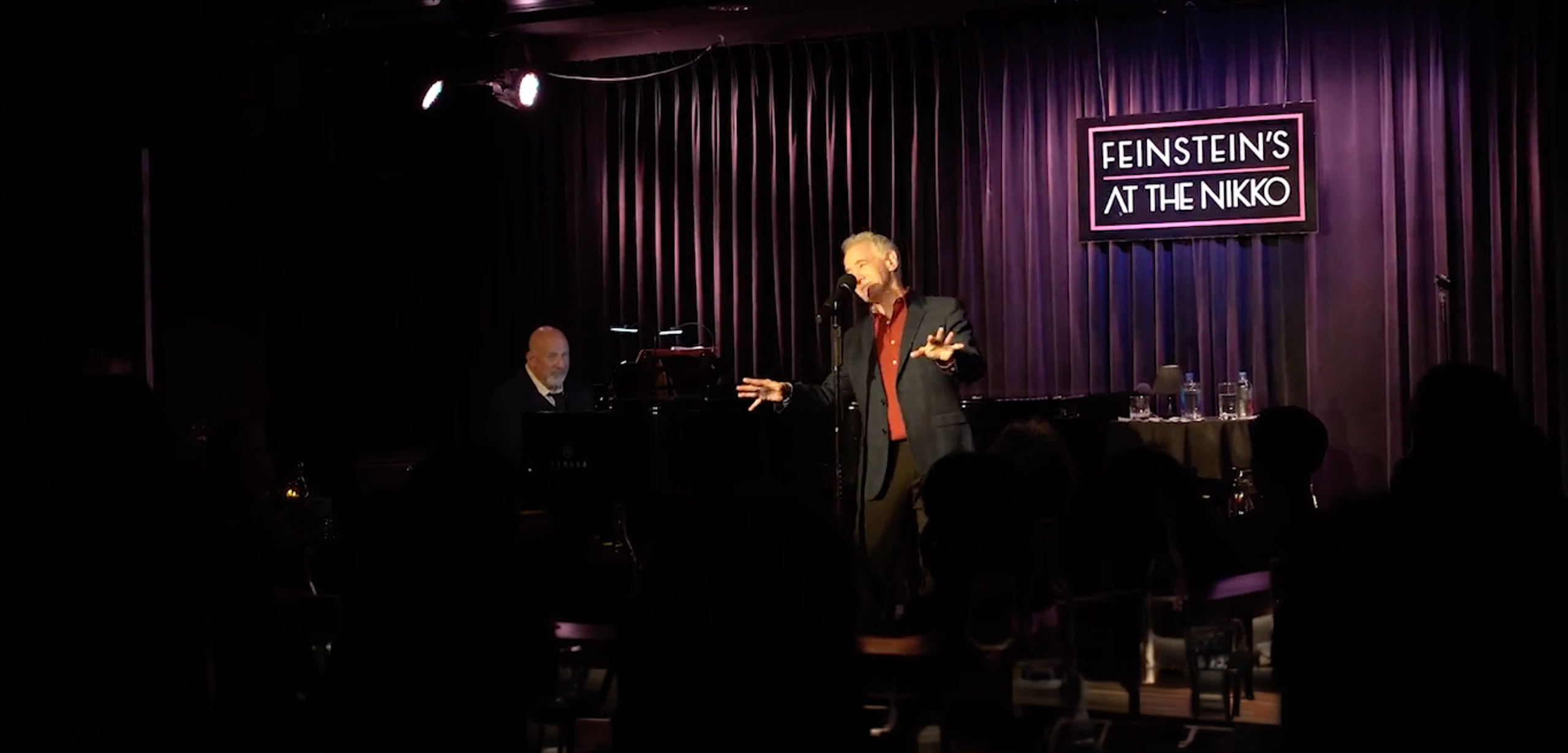 Man singing at microphone on stage with pianist in background at Feinstein's at the Nikko club.