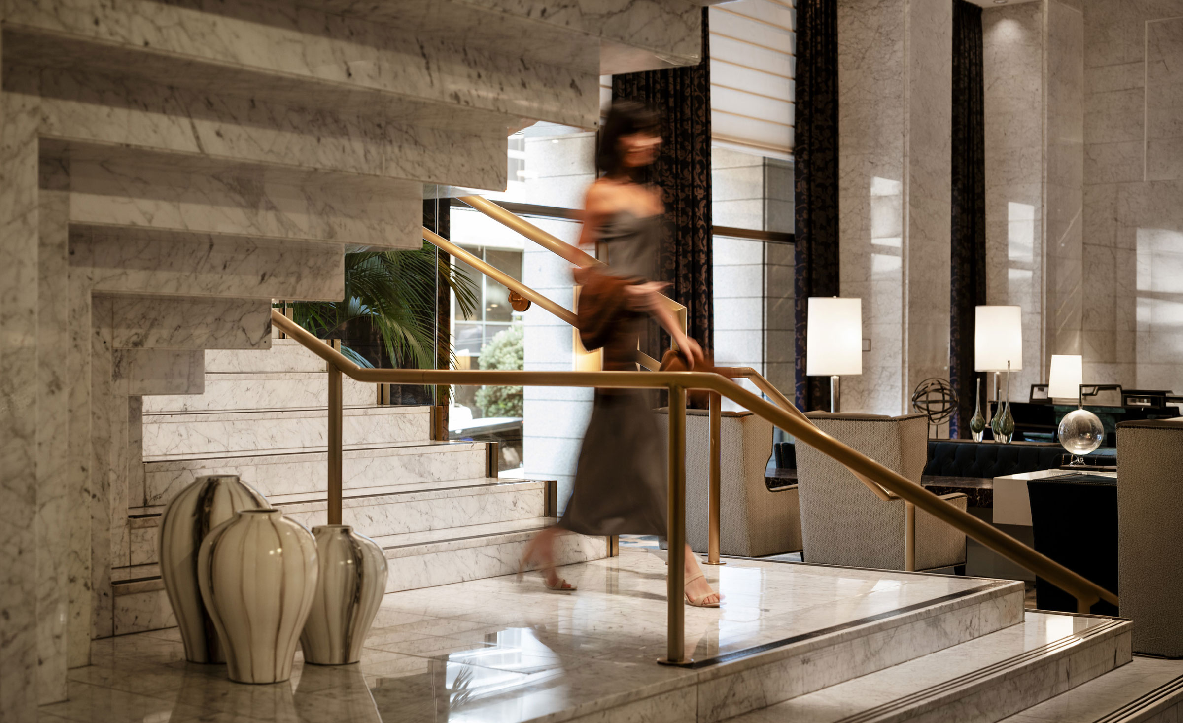 Blurred woman in a dress walking down marble stairs in a stylish, modern hotel lobby.