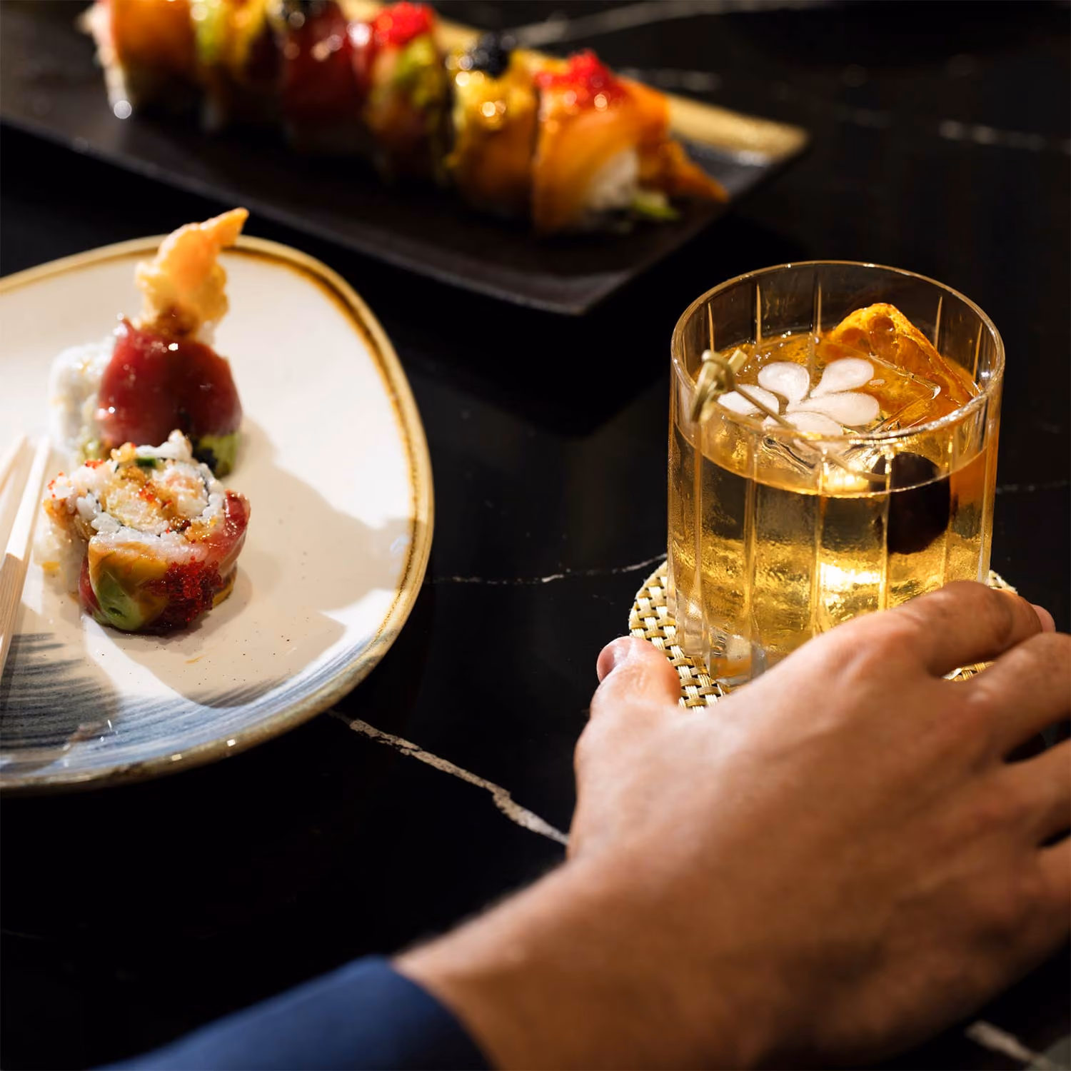 Hand reaching for a cocktail with ice, cherry, and garnish beside plates of sushi on a black table.