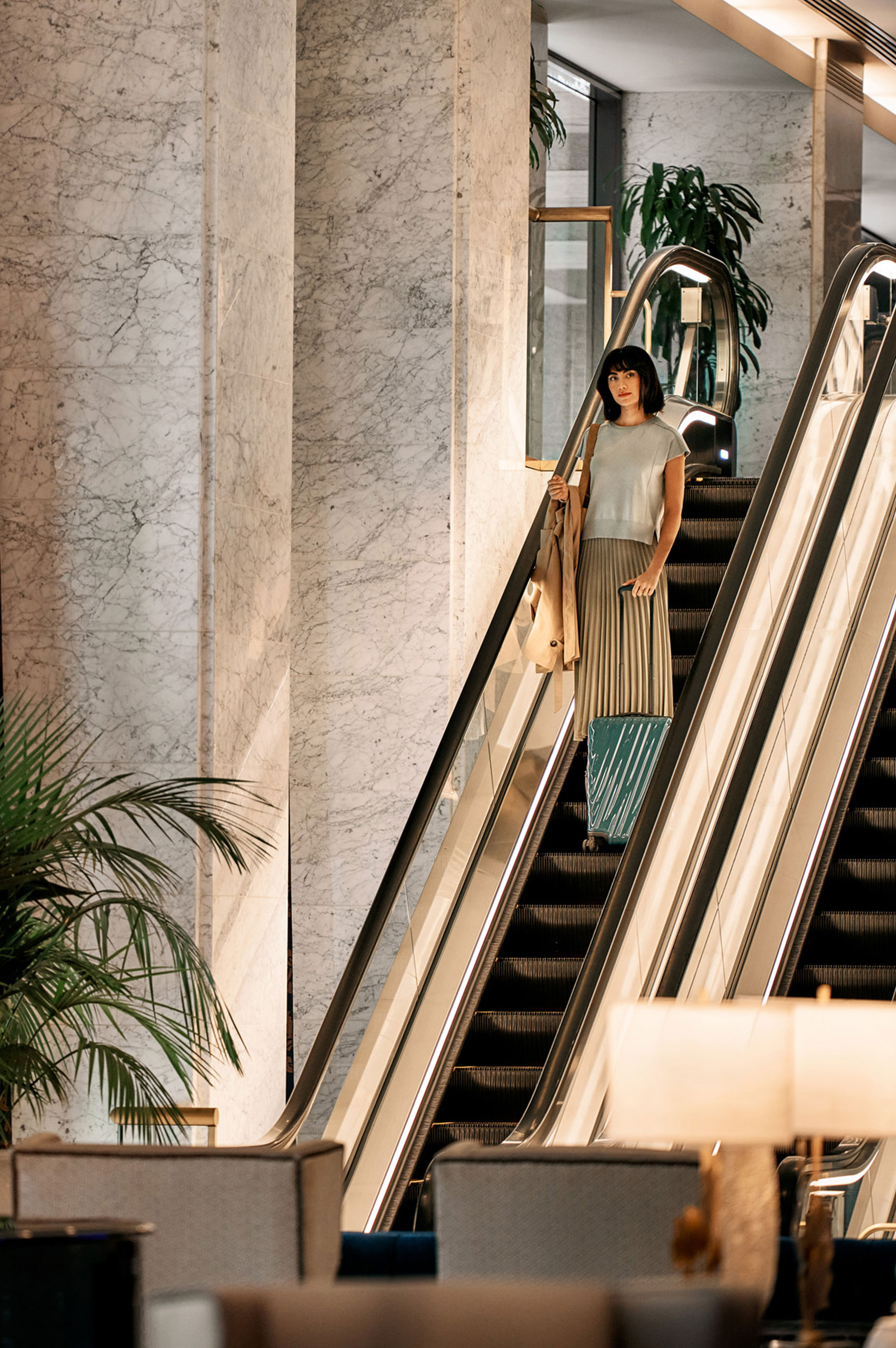 Woman in neutral outfit with suitcase rides down escalator in marble-walled hotel lobby.