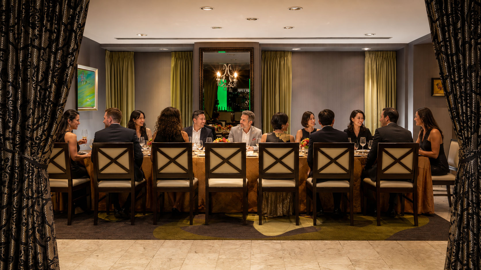 Group of guests in formal attire seated around a long table, dining and socializing in an elegant private dining room.