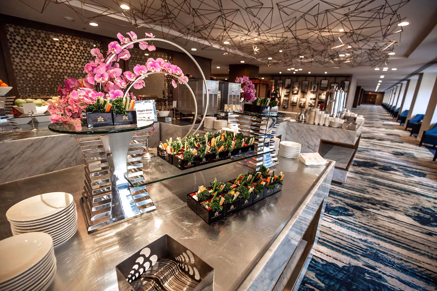 Buffet table with trays of veggie sticks, plates, and pink orchid decorations in a modern dining area.