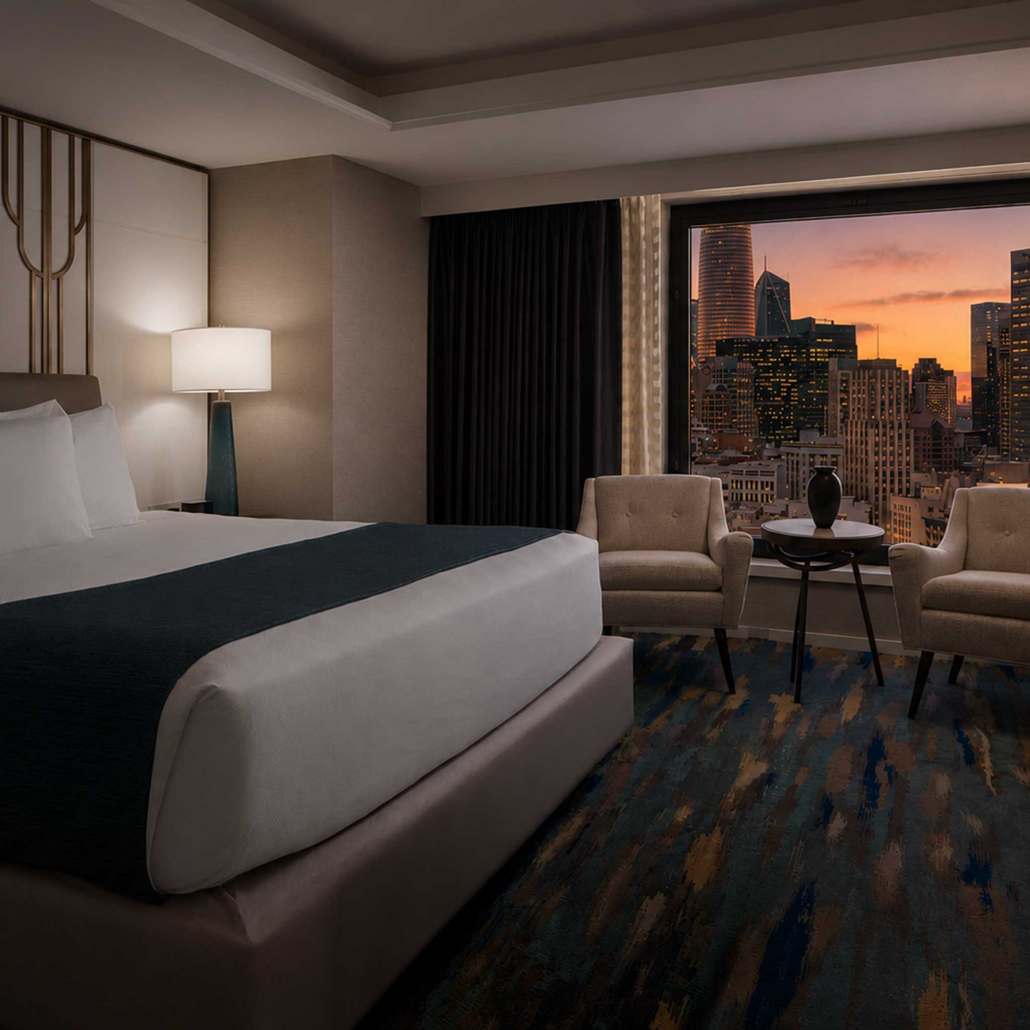 Hotel room with a king bed, two armchairs, a side table, and a city view at sunset through the window.