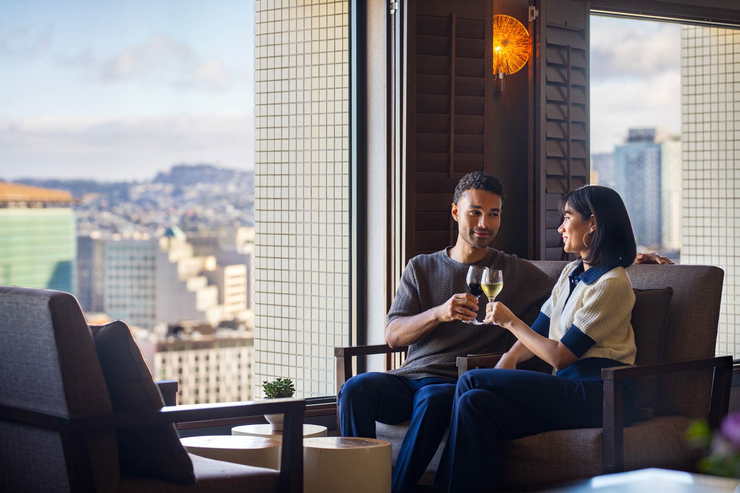 Couple sitting in a lounge by a window overlooking a cityscape, toasting with red and white wine glasses.