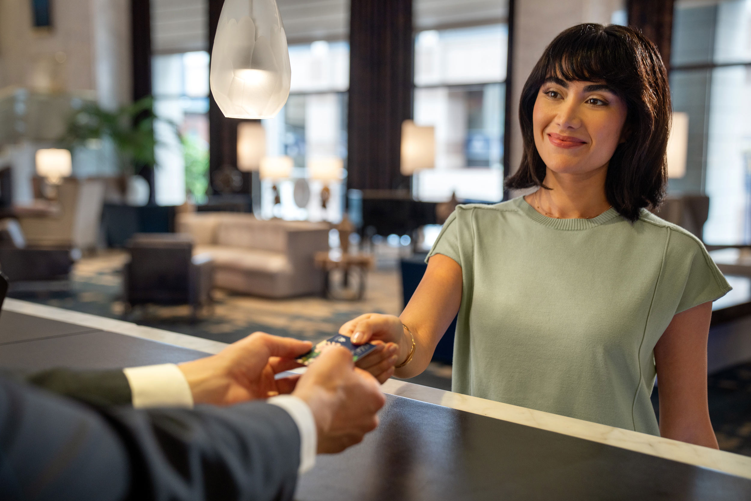Smiling woman handing a card to a person at a hotel front desk with a blurred lounge in the background.