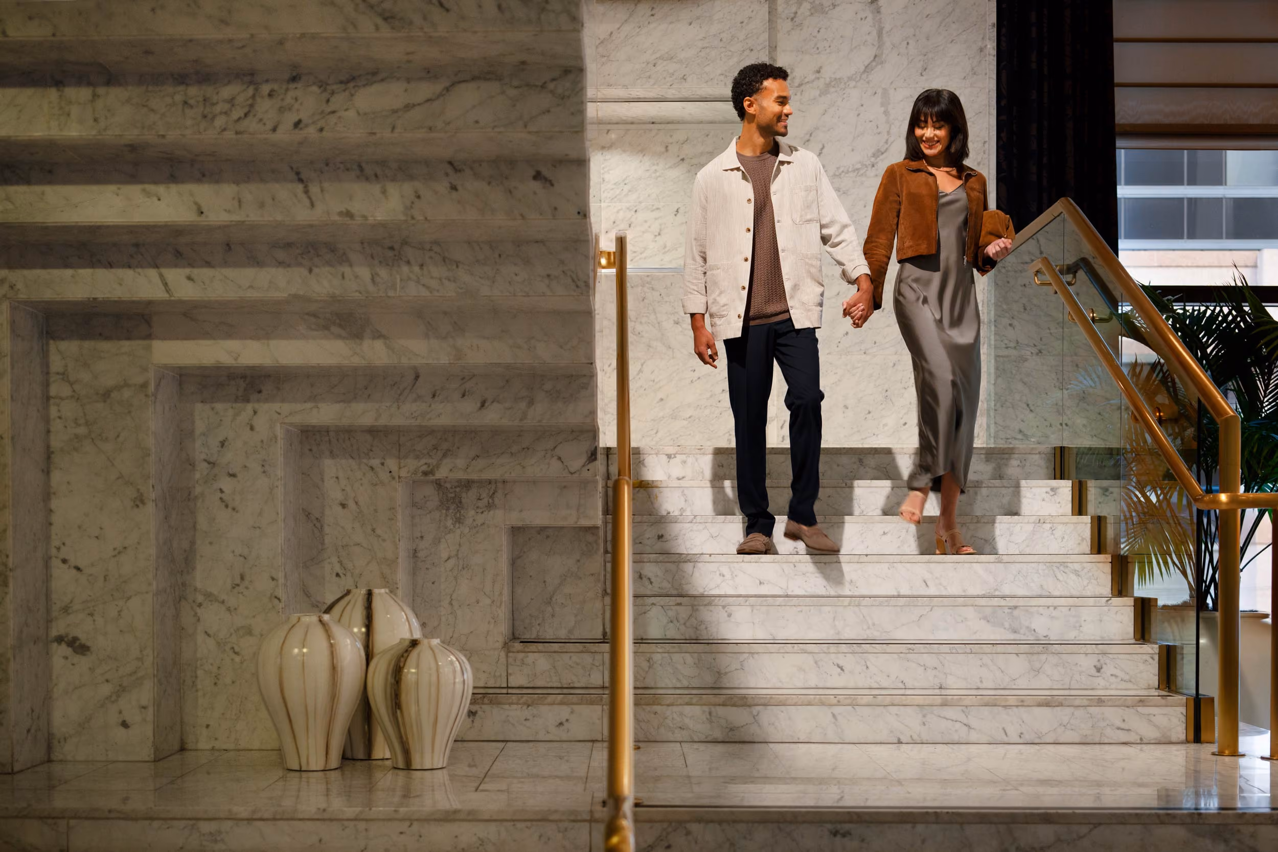 Couple holding hands and descending marble stairs in a modern indoor space with decorative vases.