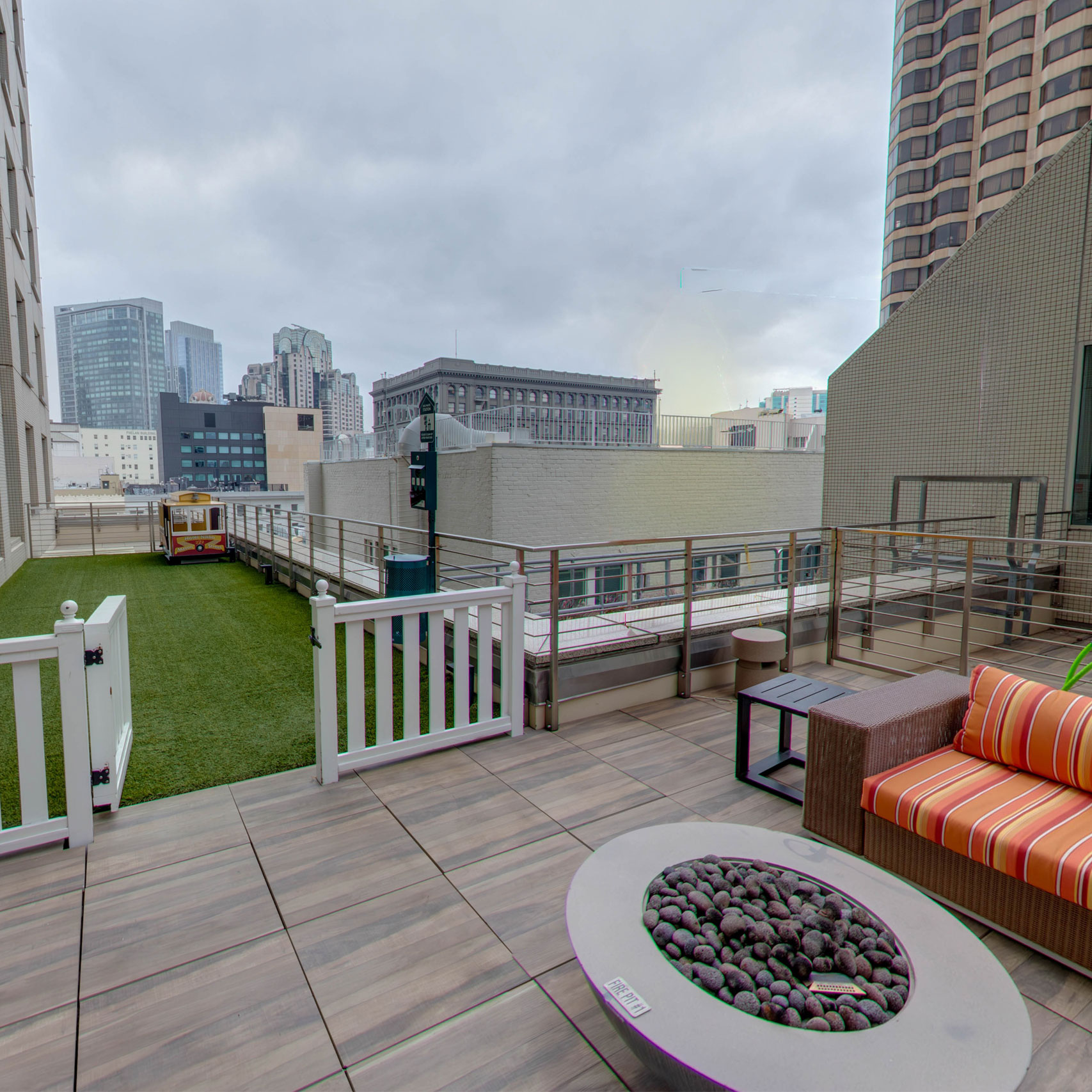 Urban rooftop patio with fire pit, striped sofa, artificial grass, and city skyline under cloudy sky.