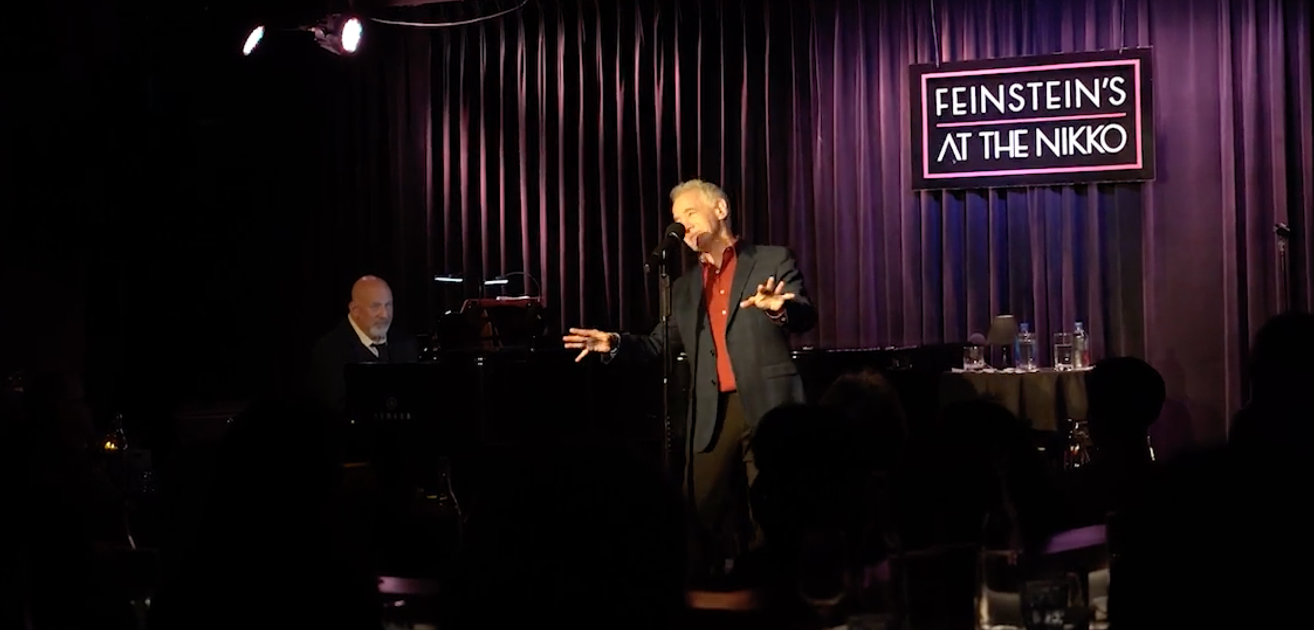 A man in a dark suit and red shirt singing into a microphone on stage with a pianist playing a grand piano in the background, and a sign on the wall reading 'FEINSTEIN'S AT THE NIKKO', with a small table holding water bottles and glasses on the right, and an audience in front in dim lighting.