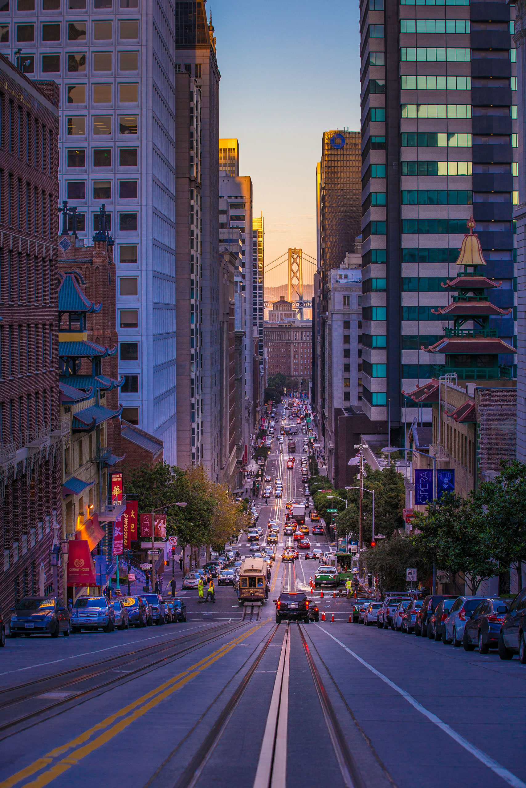 San Francisco street with cable car tracks, tall buildings, cars, trees, and Bay Bridge in the distance at sunset.