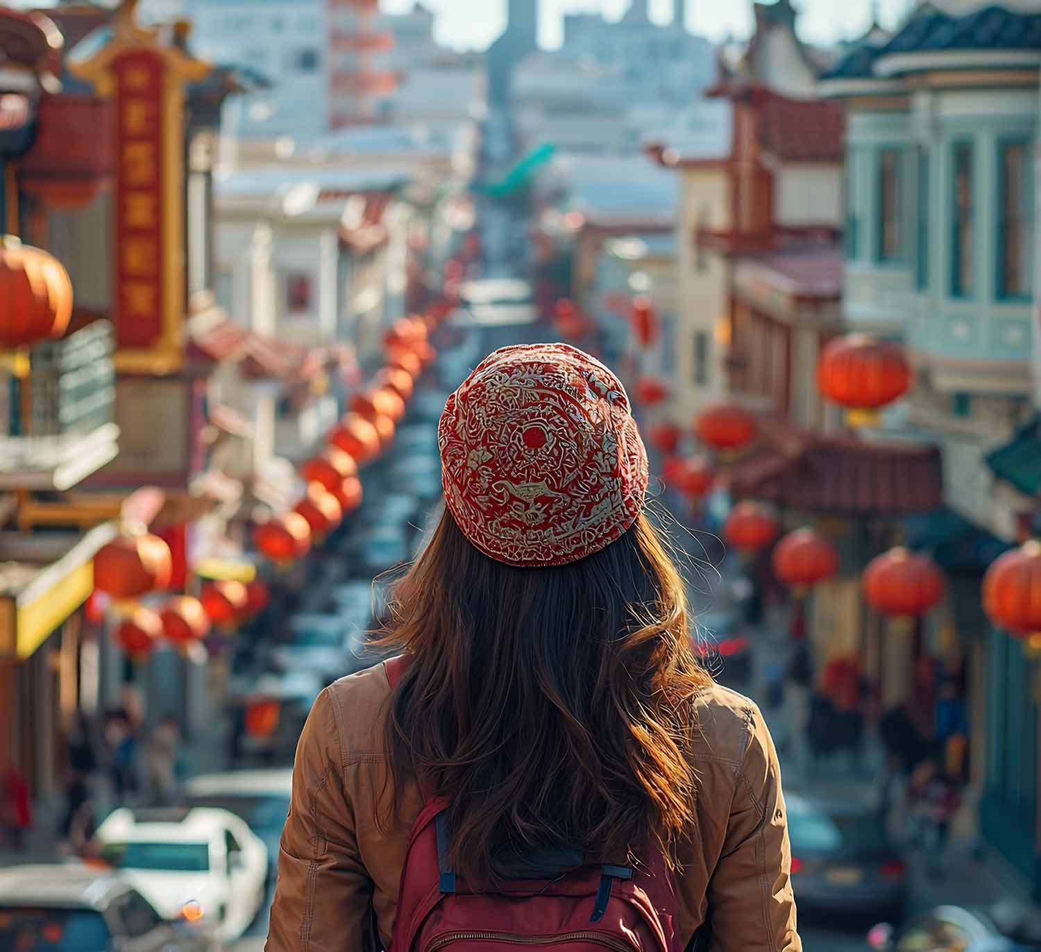 Person with long hair wearing a patterned red hat and brown jacket looks down a street lined with red lanterns and buildings in a Chinatown district.