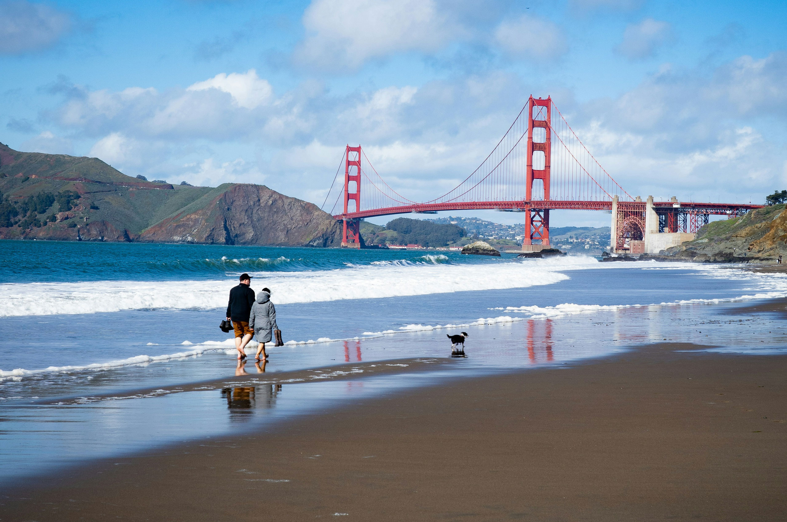 A couple walking barefoot along a sandy beach with a small black and white dog, with the red Golden Gate Bridge and green hills in the background under a partly cloudy blue sky.