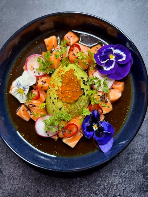 Top view of a gourmet dish featuring diced salmon sashimi arranged around a mound of green avocado cream topped with bright orange salmon roe, garnished with thin radish slices, red chili slices, edible flowers in purple and white, green herbs, and black sesame seeds, all served in a shallow black bowl with a dark broth.