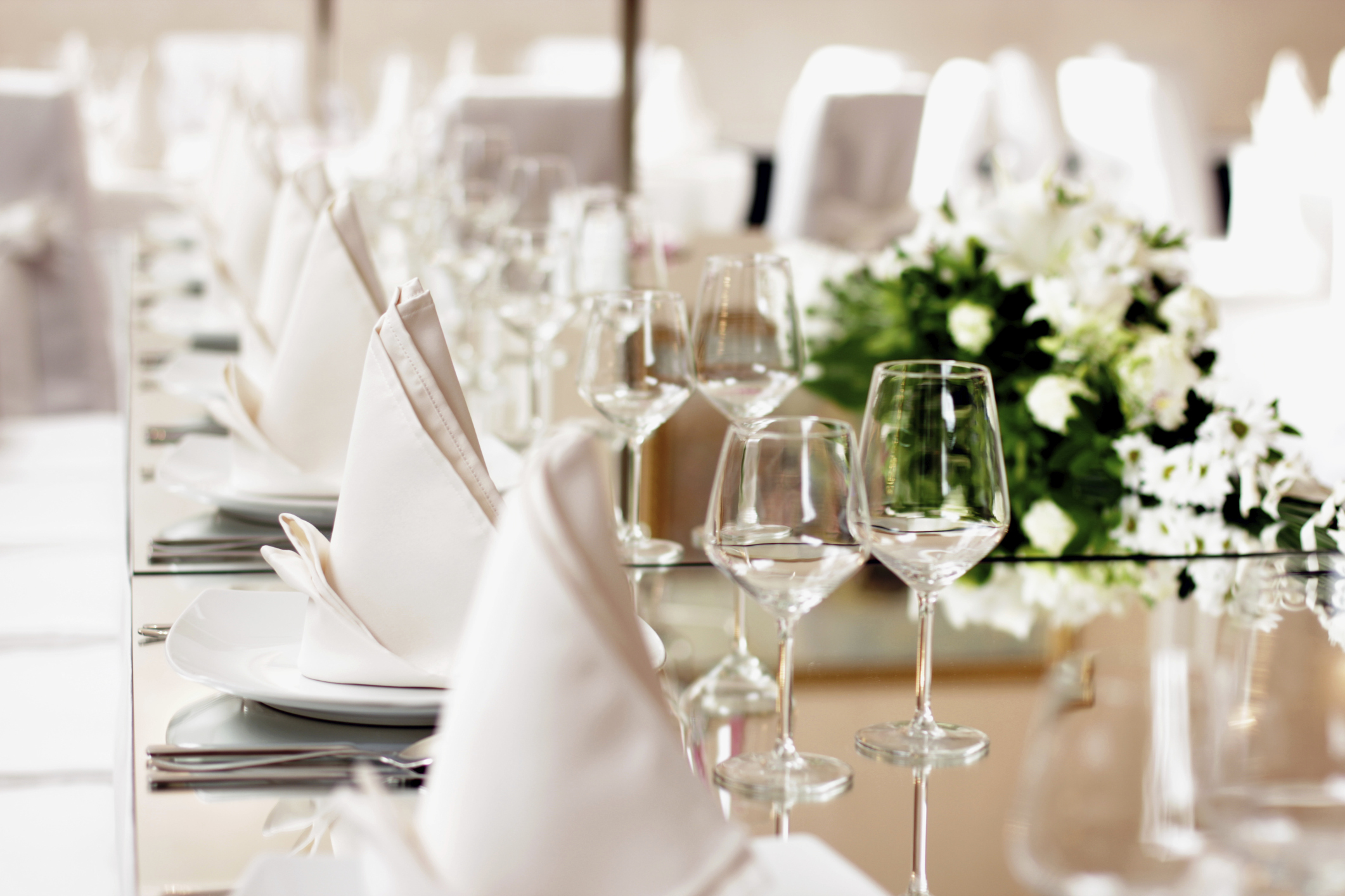 Elegant dining table set with white folded napkins on plates, clear wine glasses, silverware, and a floral centerpiece with white flowers and greenery.