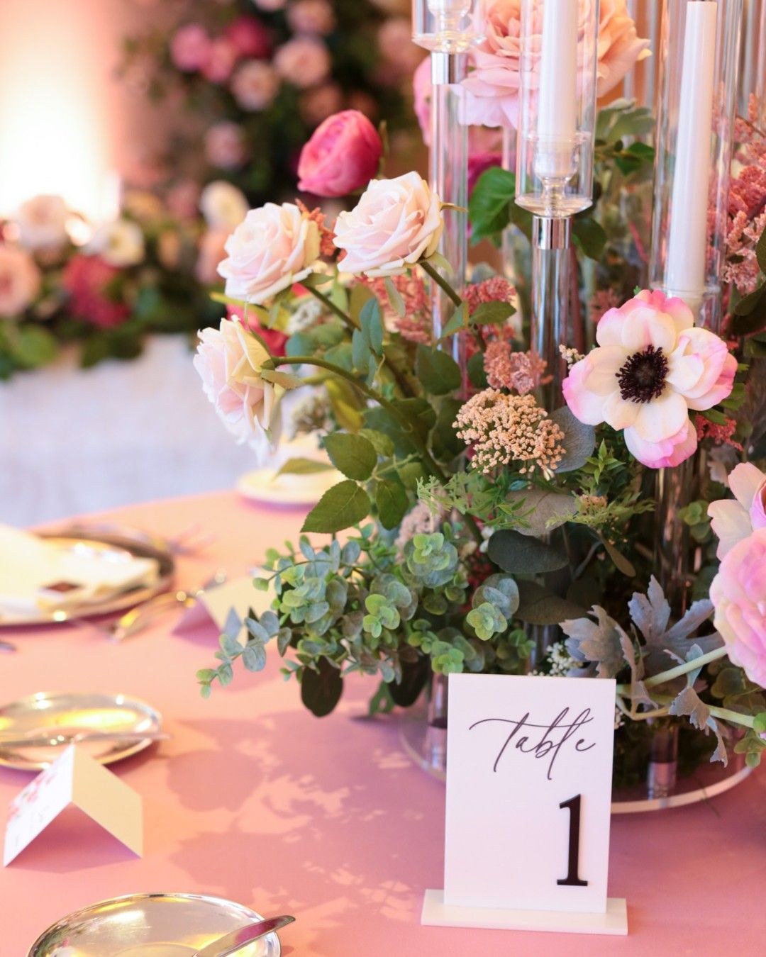 A floral centerpiece featuring pink and white roses, eucalyptus, and other greenery arranged around tall glass candle holders on a pink tablecloth with a white card labeled 'Table 1'.