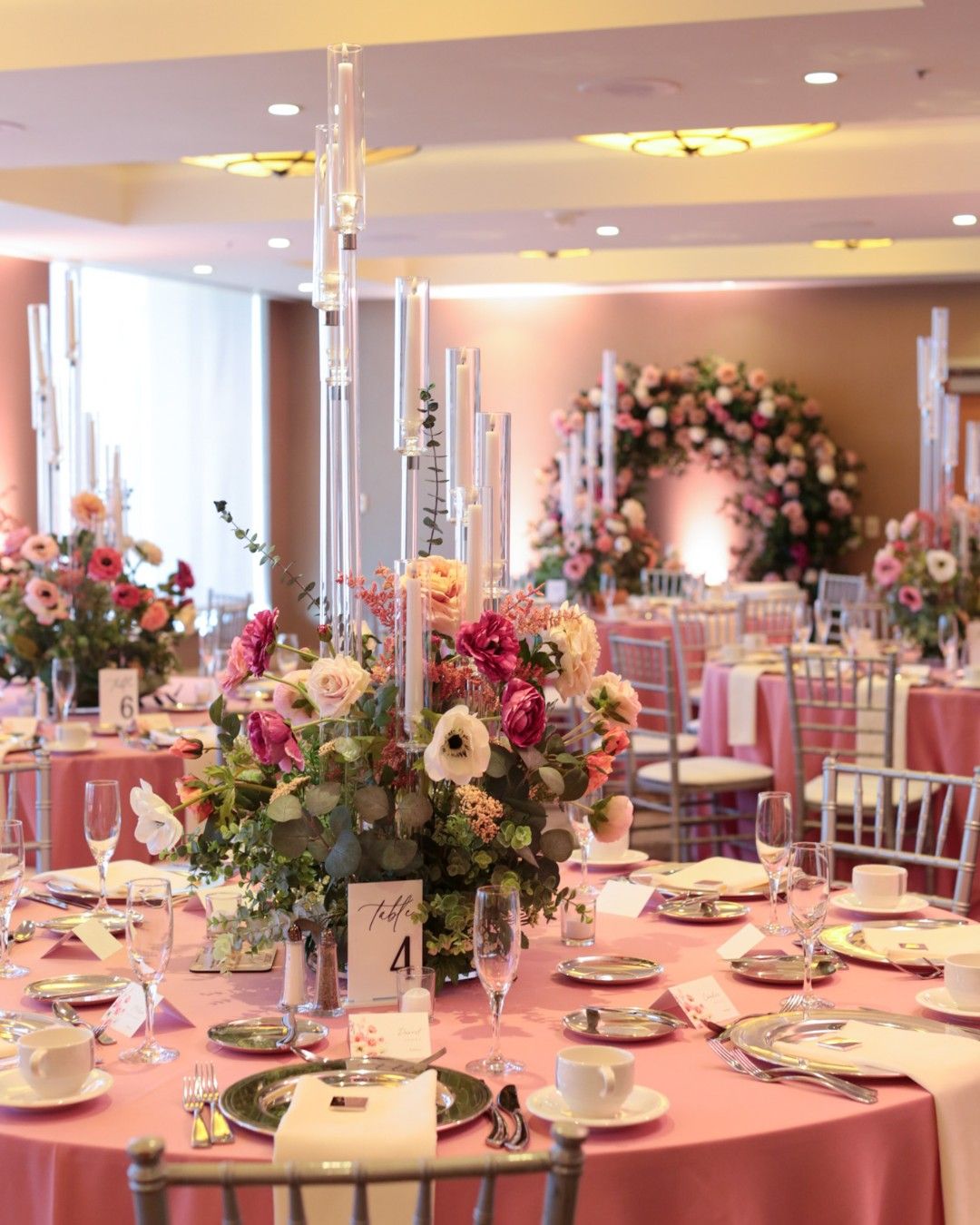 Elegant wedding reception tables with pink tablecloths, silver chairs, and floral centerpieces featuring pink and white flowers with tall clear candle holders, arranged around fine dinnerware including plates, silverware, glasses, and white cups, with a floral arch decorated with similar flowers in the background.
