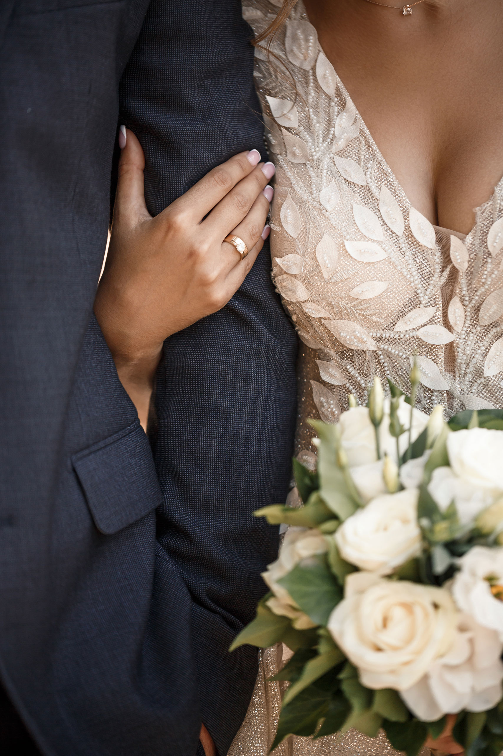Bride's arm with a wedding ring holding the groom's arm, the bride is wearing a detailed lace dress with floral patterns and holding a bouquet of white roses and green leaves.