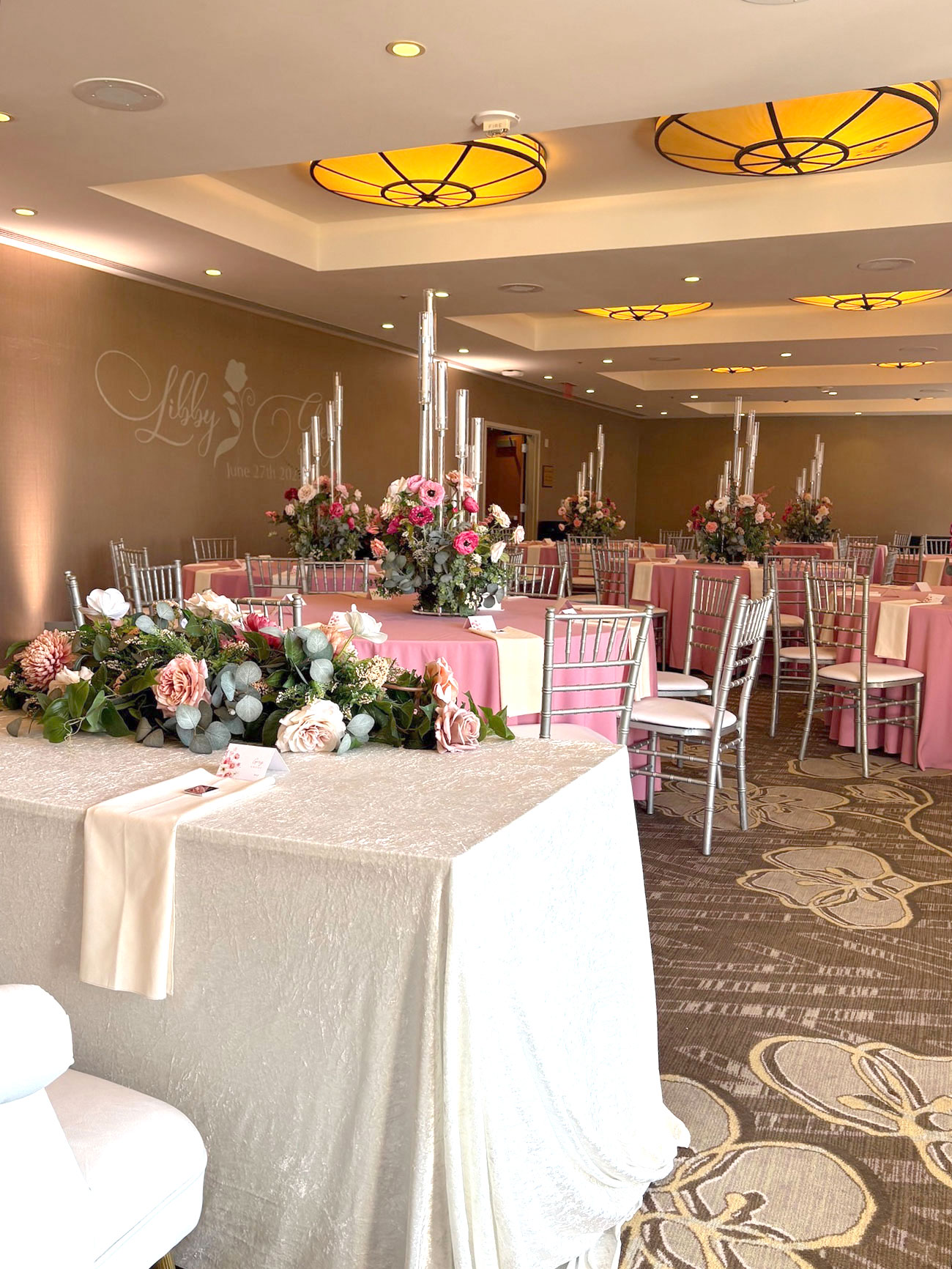 Indoor event space set up for a celebration with pink and beige tablecloths, silver chairs, floral centerpieces with pink flowers, and tall candle holders, featuring a beige table with a floral arrangement in the foreground.