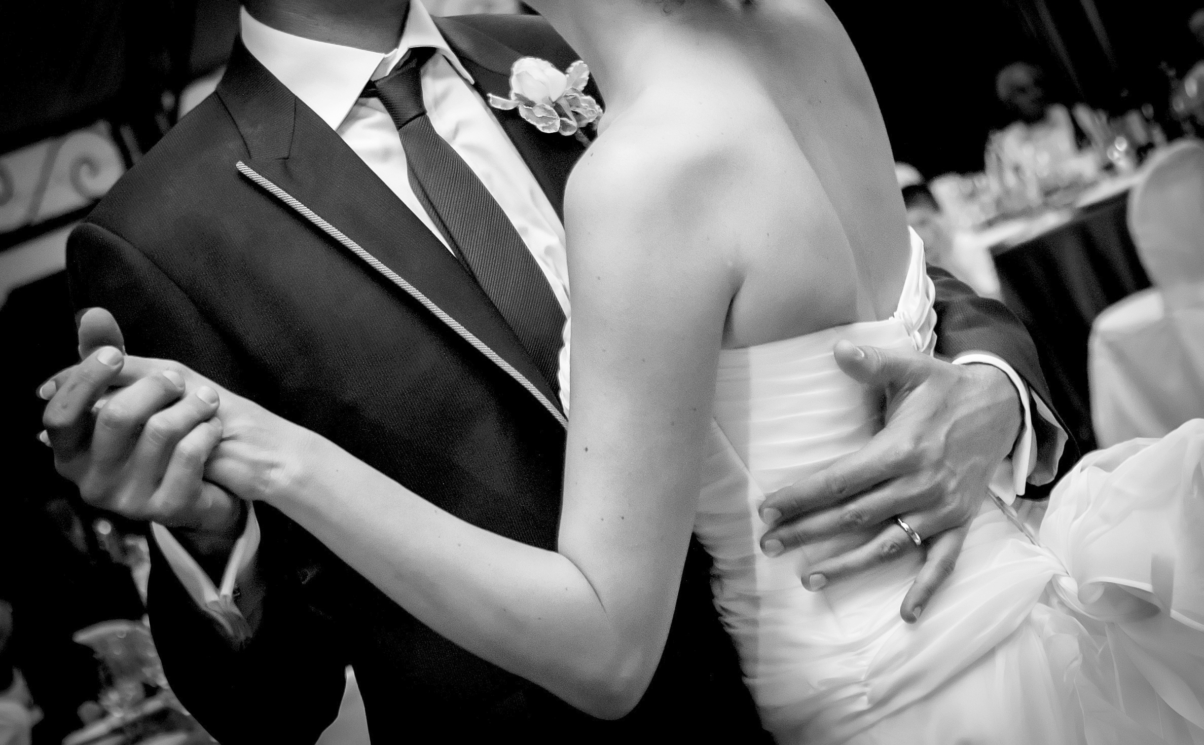 Black and white close-up of a bride and groom dancing, showing their hands clasped and the groom's hand on the bride's back, with the bride wearing a strapless gown and the groom in a suit with a tie and boutonniere.