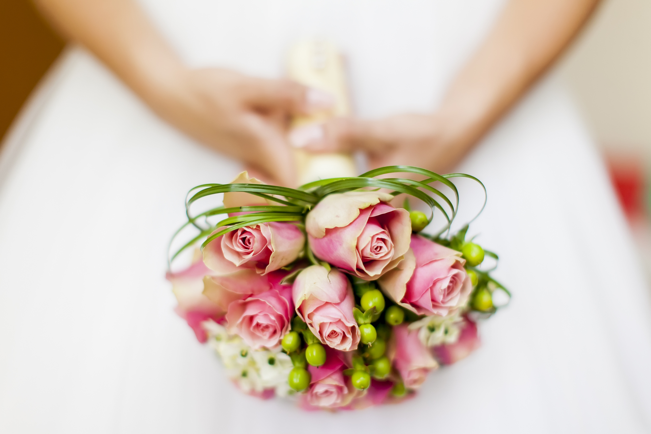 Close-up of hands holding a bouquet of pink roses with green berries and decorative grass loops, against a white blurred background.
