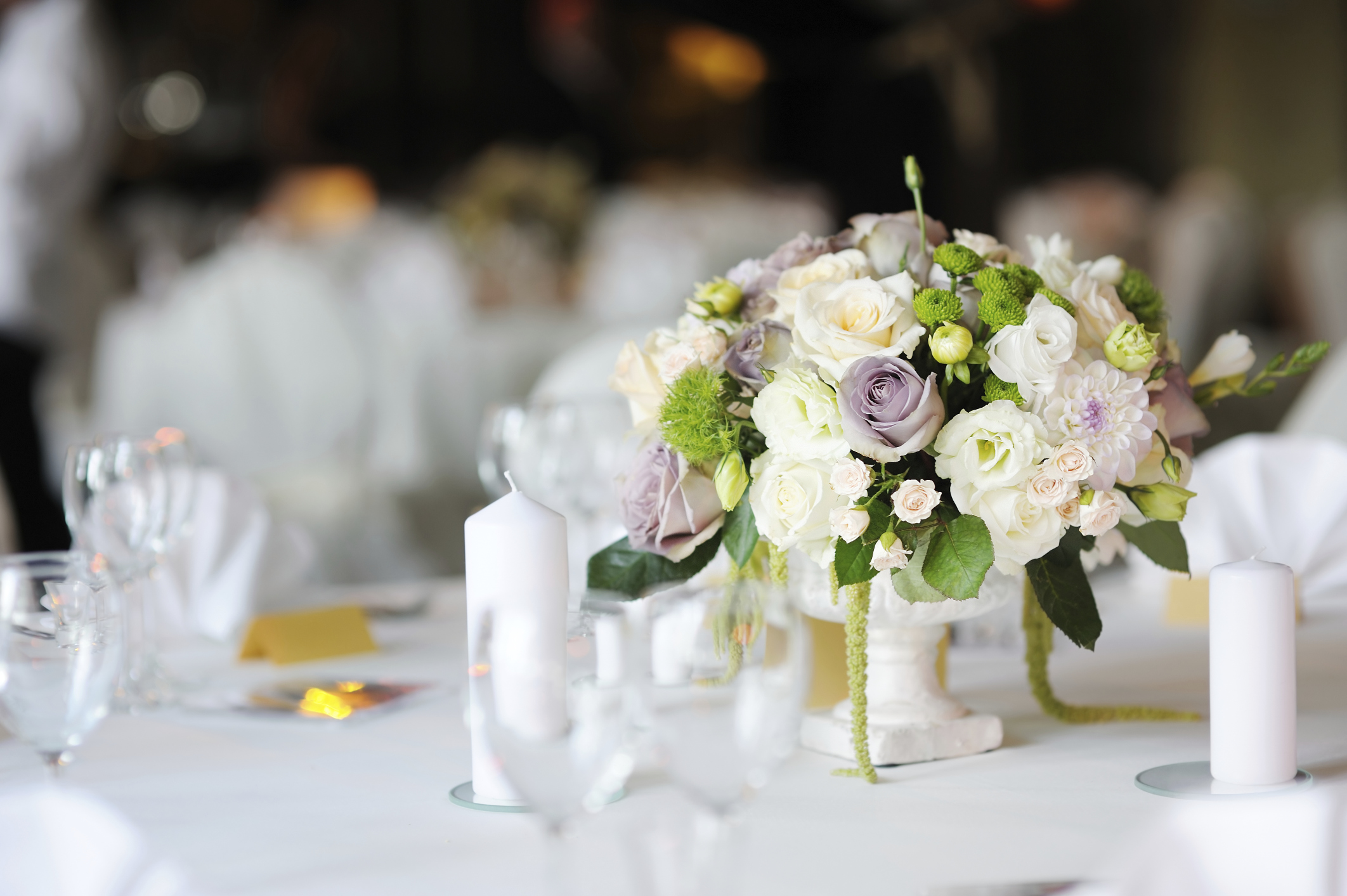Elegant wedding table centerpiece with a floral arrangement of white, pale purple, and green flowers in a white vase, surrounded by white candles and glassware on a white tablecloth.