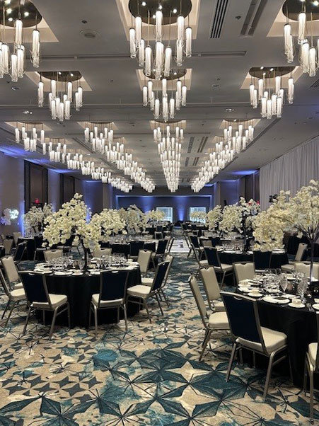Large ballroom set for an event with round tables covered in black tablecloths, each adorned with tall white floral centerpieces and place settings, under multiple modern chandeliers hanging from the ceiling.