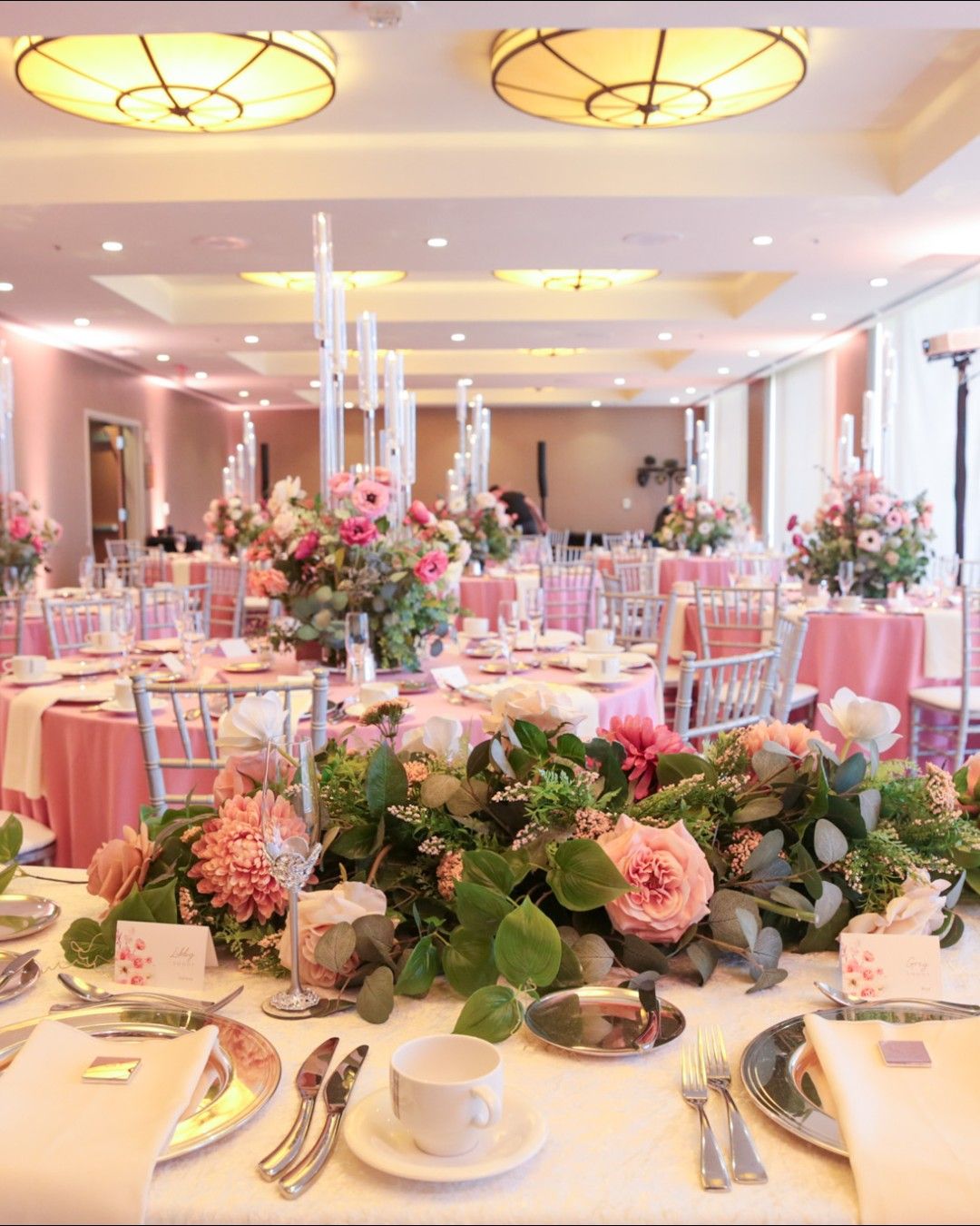 Elegant wedding reception room with round tables covered in pink tablecloths, adorned with tall floral centerpieces and candles, surrounded by silver chairs and set with white napkins, plates, and tea cups, featuring a lush floral arrangement of pink roses and greenery in the foreground.