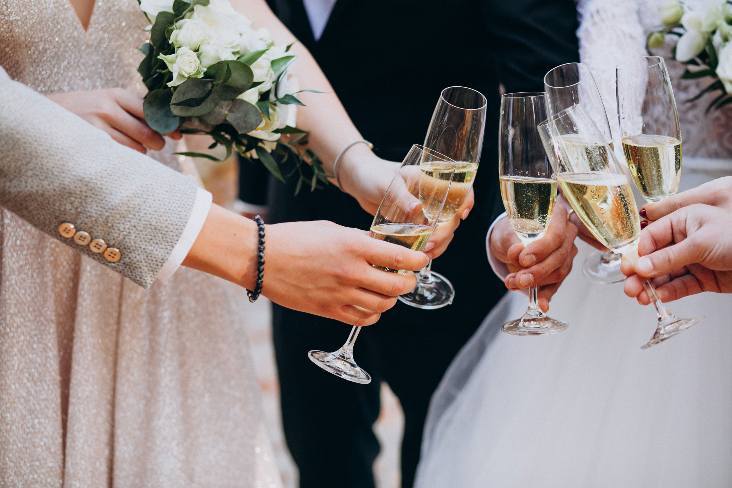 Close-up of six hands holding champagne flutes raised for a toast, with two people holding white floral bouquets, suggesting a wedding celebration.