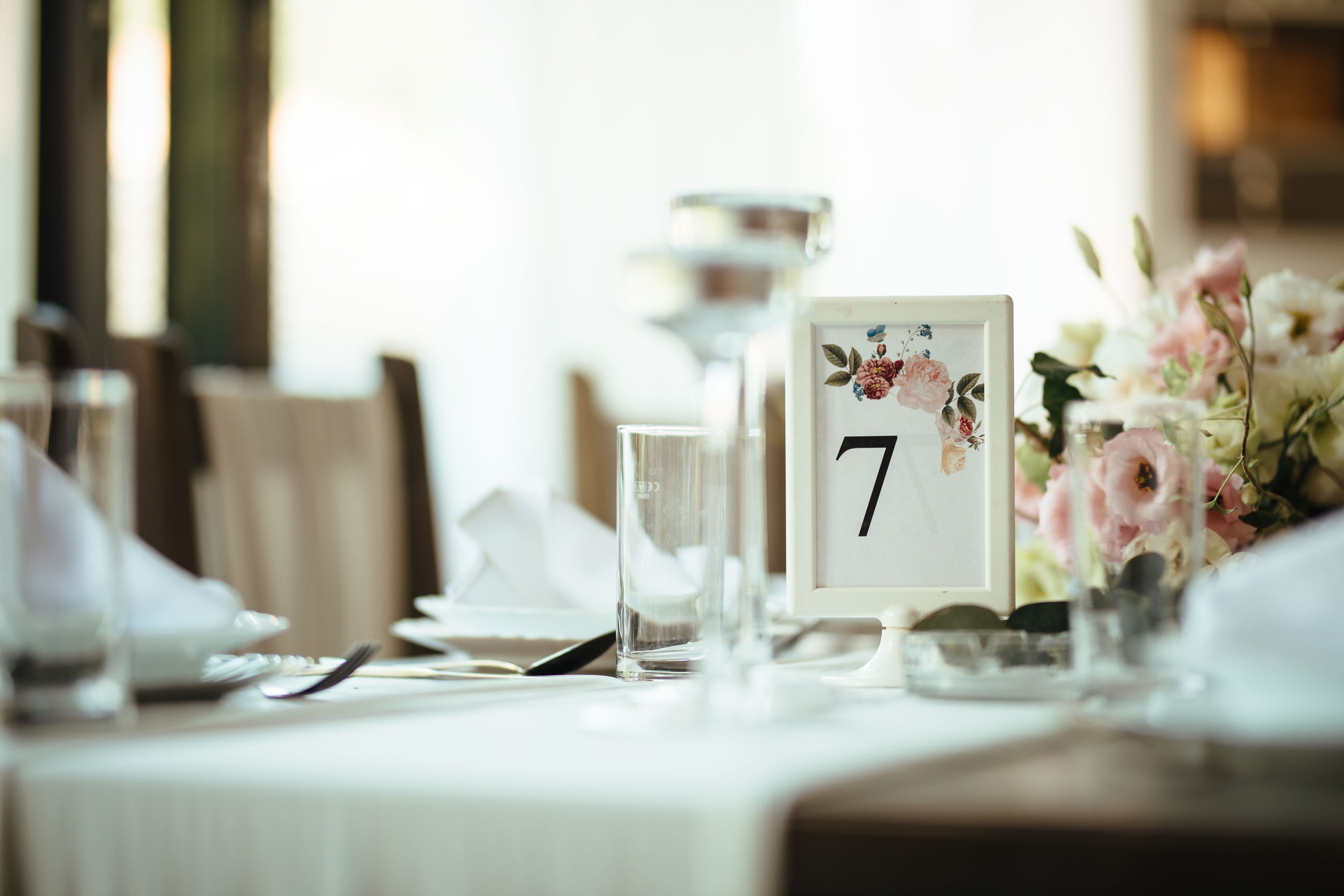 A decorated wedding reception table with a white tablecloth, glassware, folded napkins, and a floral centerpiece, featuring a table number 7 sign adorned with flowers.