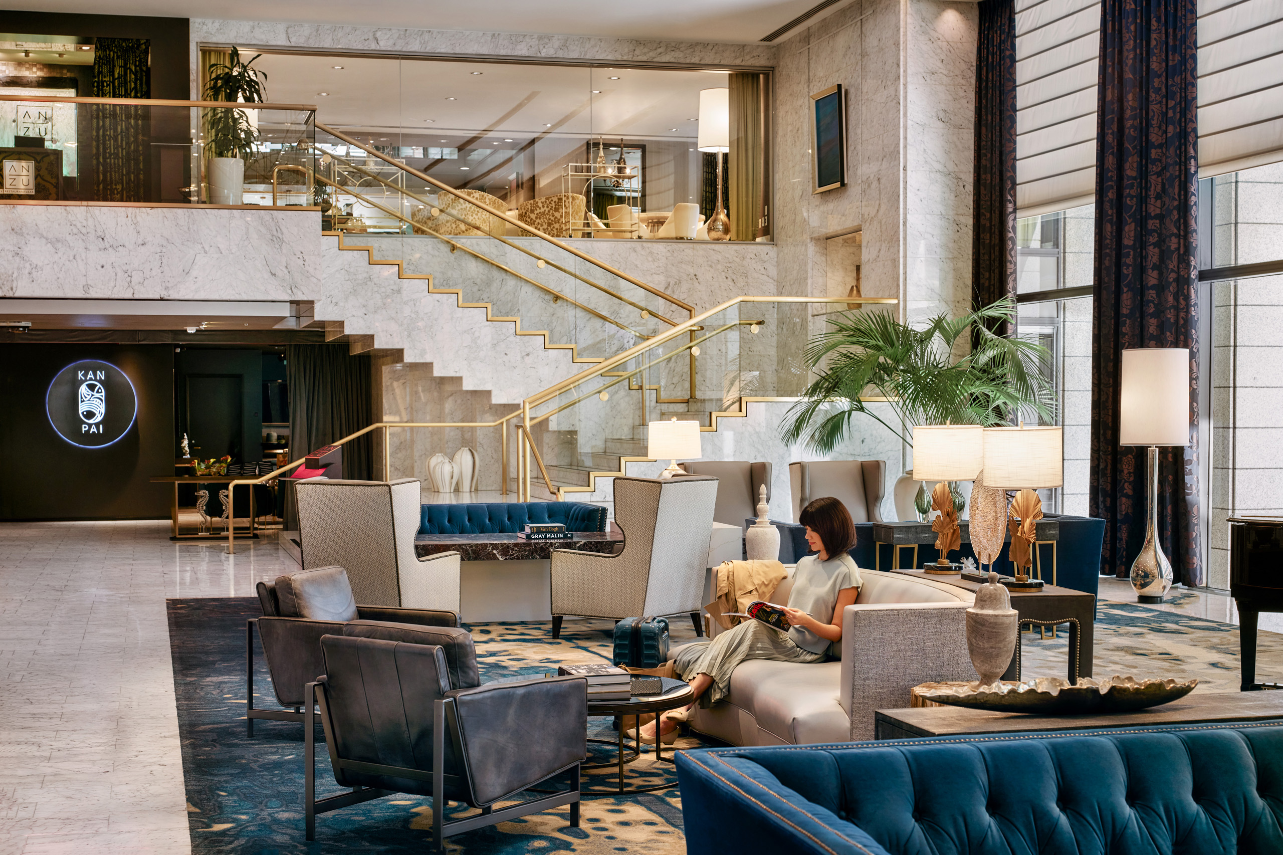 Modern hotel lobby with a woman sitting on a light gray sofa reading a magazine, surrounded by various armchairs and sofas, a large potted plant, lit table lamps, and a marble staircase with gold railings in the background.