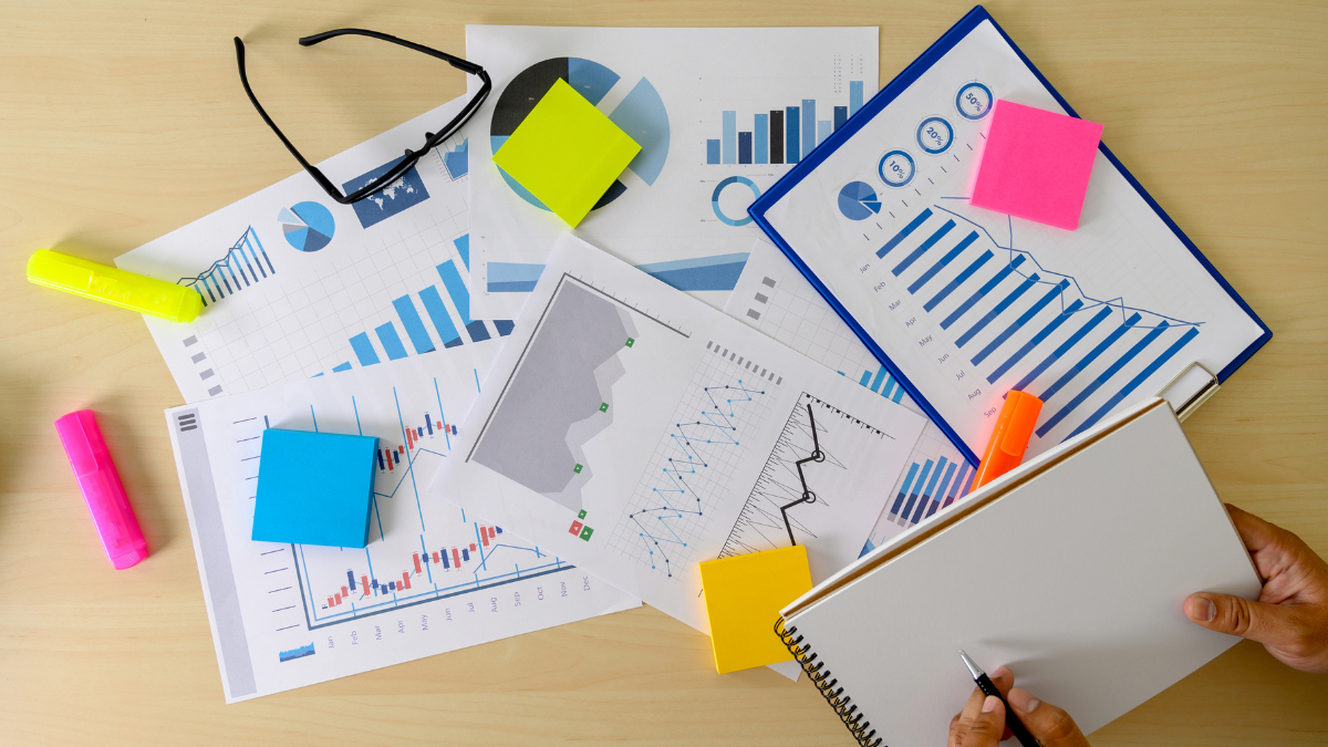A desk with various financial charts, graphs, sticky notes, highlighters, glasses, and a notebook with a pen.