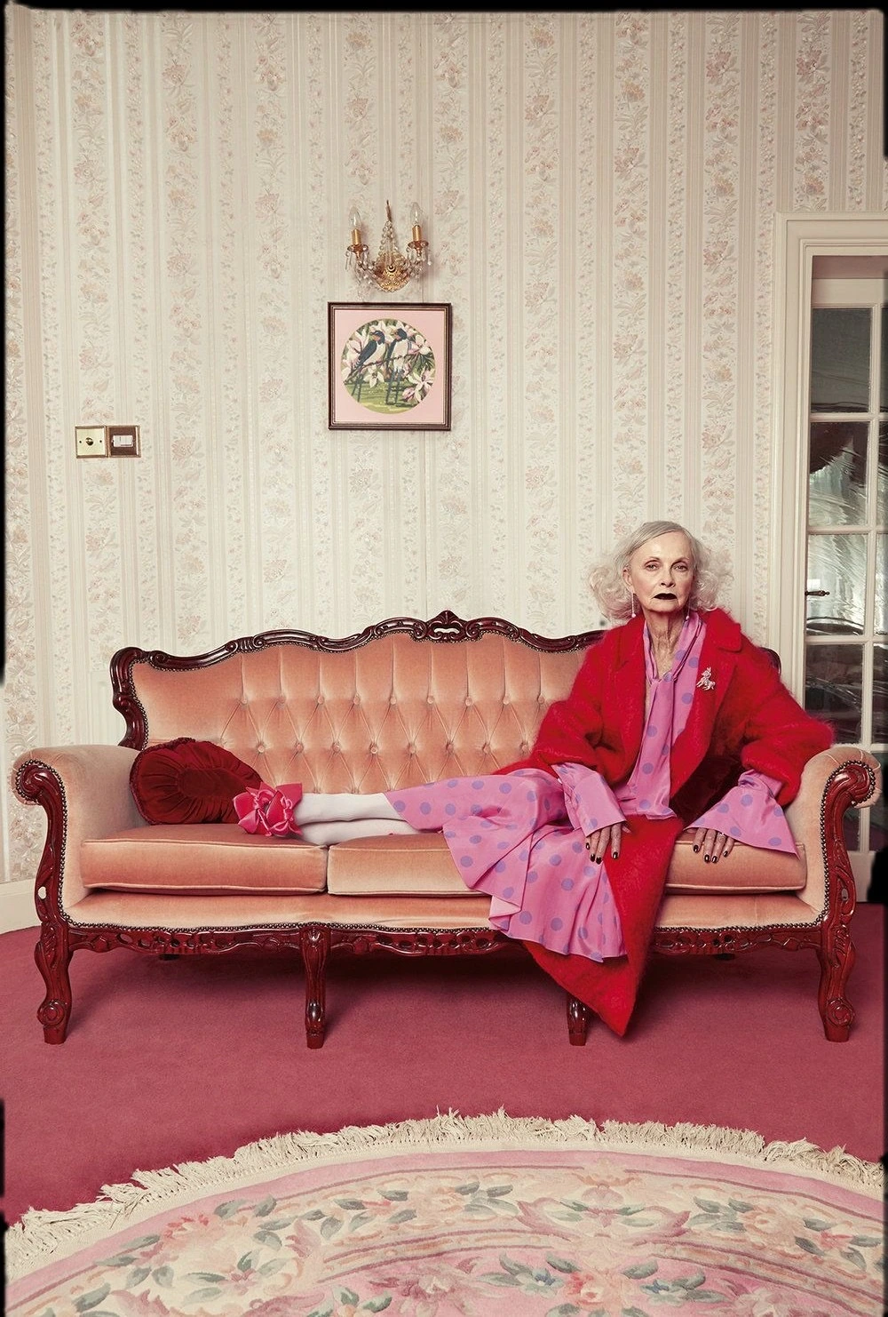 An older woman in a red coat seated on a pink sofa in a softly patterned room