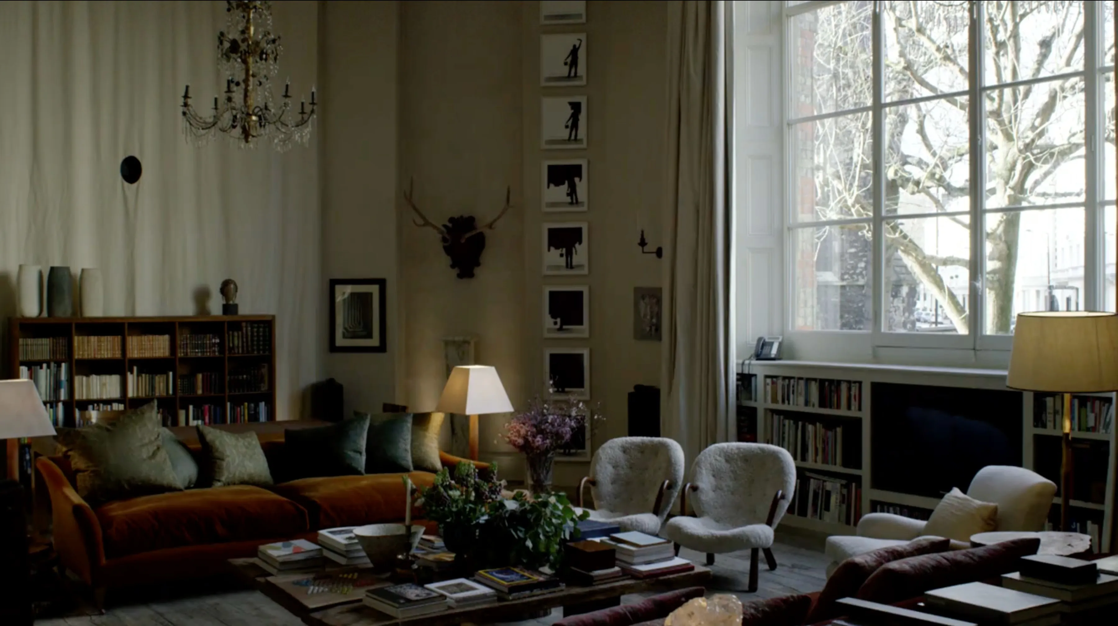 A sitting room with a velvet sofa, white armchairs, shelves of books and a wall of framed prints, lit by lamps beside tall windows