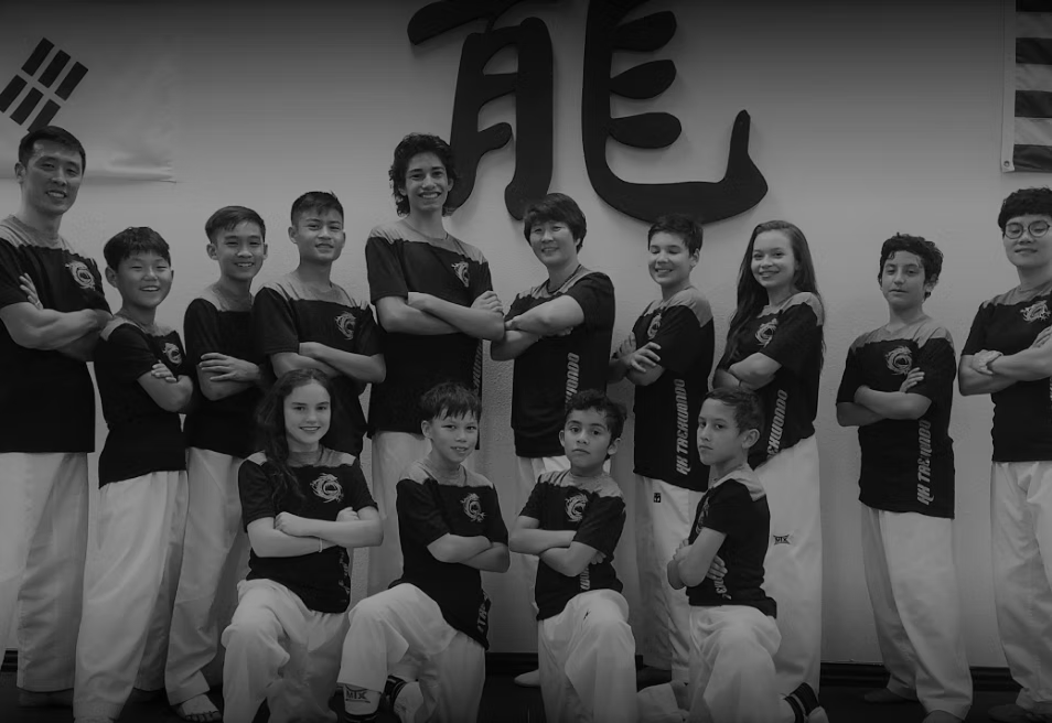 Group of young Taekwondo students in uniform posing with arms crossed inside a martial arts studio.