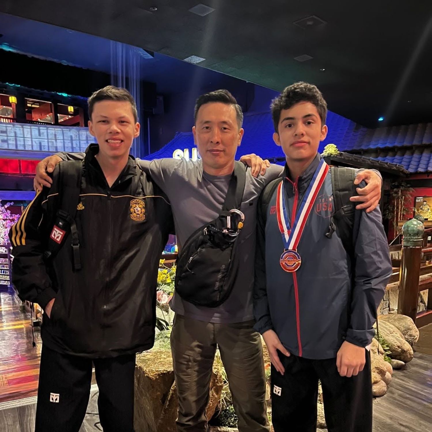 Three males posing indoors, the one on the right wearing a medal around his neck and all wearing backpacks.