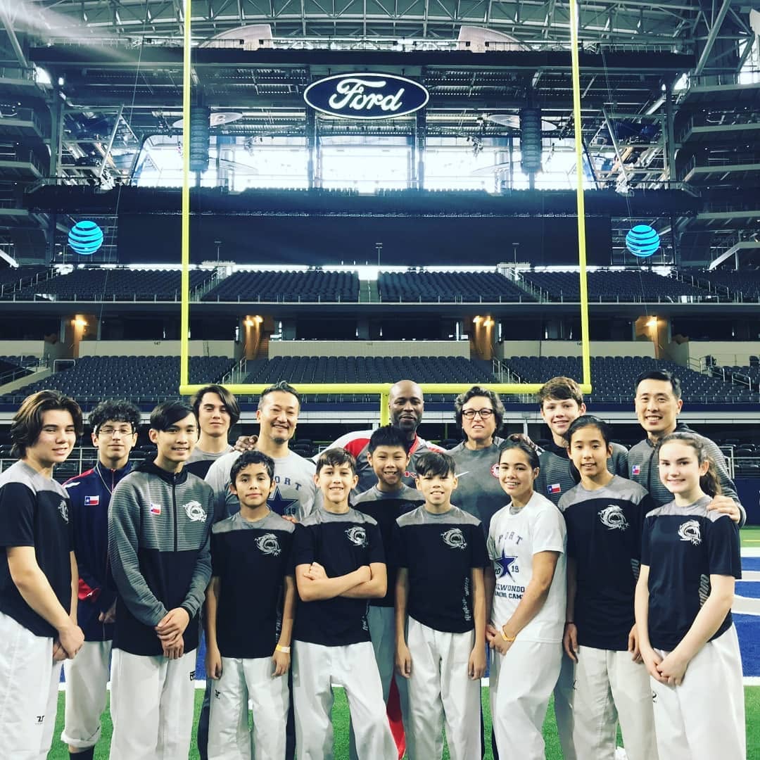 Group of young martial artists and coaches posing on a football field inside a stadium under a Ford sign.