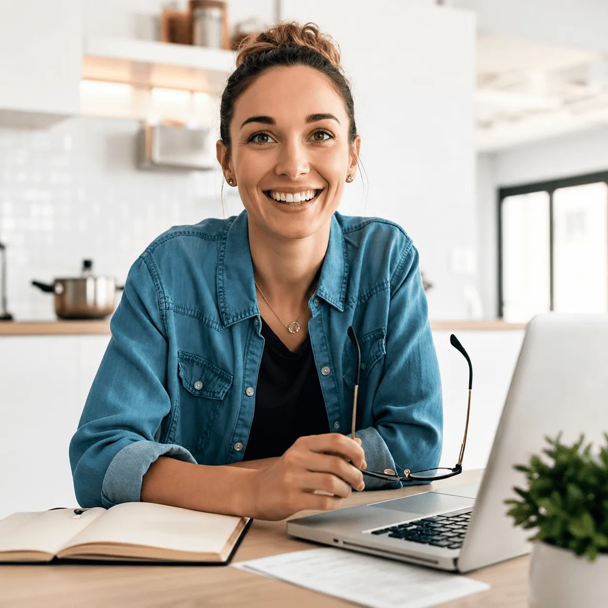 Smiling woman in denim shirt holding glasses at desk with laptop, notebook, and plant.