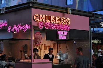 Food stand with neon sign reading 'CHURROS & Chocolate' and people ordering inside.