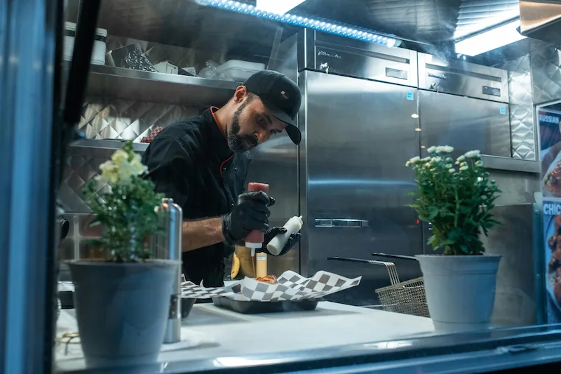 Chef in black uniform adds sauce to food on a tray inside a commercial kitchen.