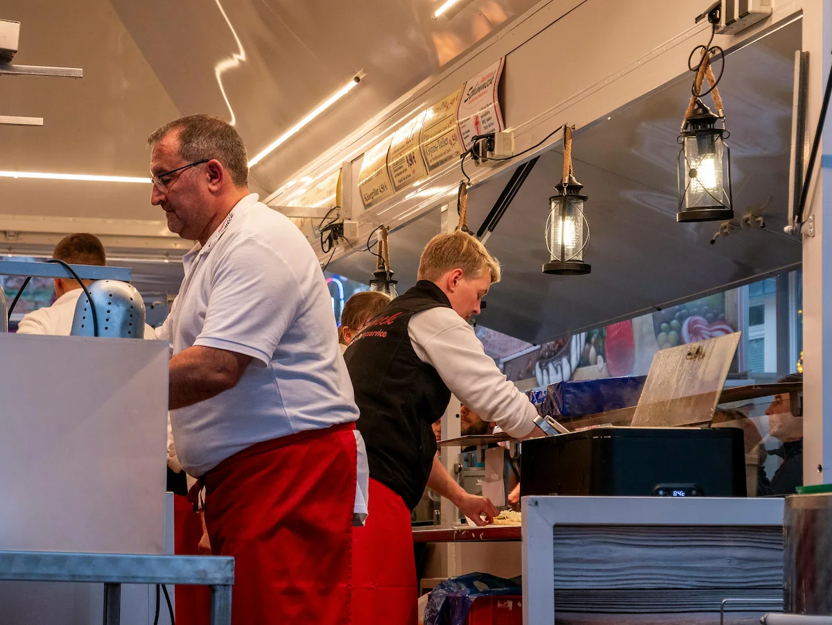 Two people wearing red aprons preparing food in a well-lit kitchen or food stall.
