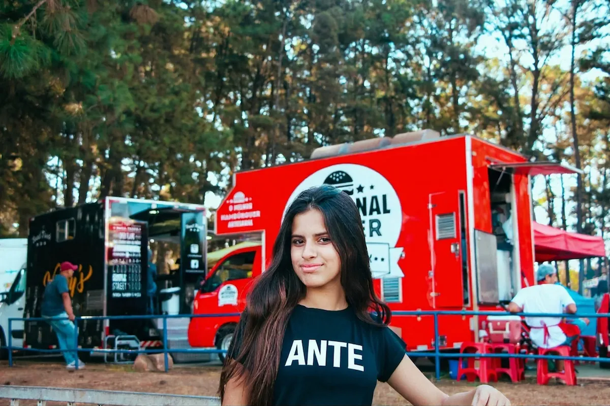 Young woman with long dark hair wearing a black shirt with 'ANTE' stands outdoors near red and black food trucks.