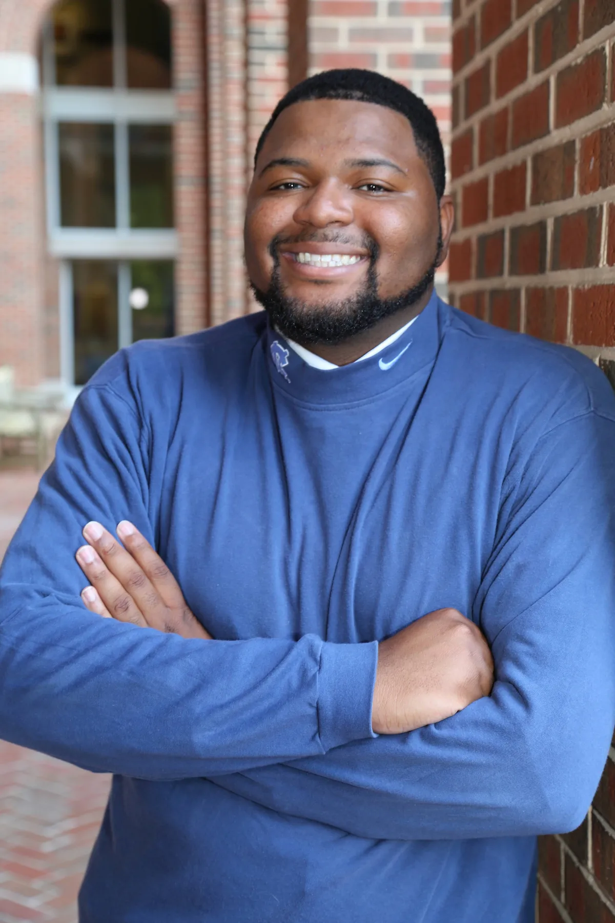 Smiling man in a blue long-sleeve shirt stands with arms crossed against a brick wall.