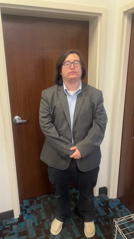 Young person wearing glasses, gray blazer, blue shirt, standing in front of a closed wooden door.