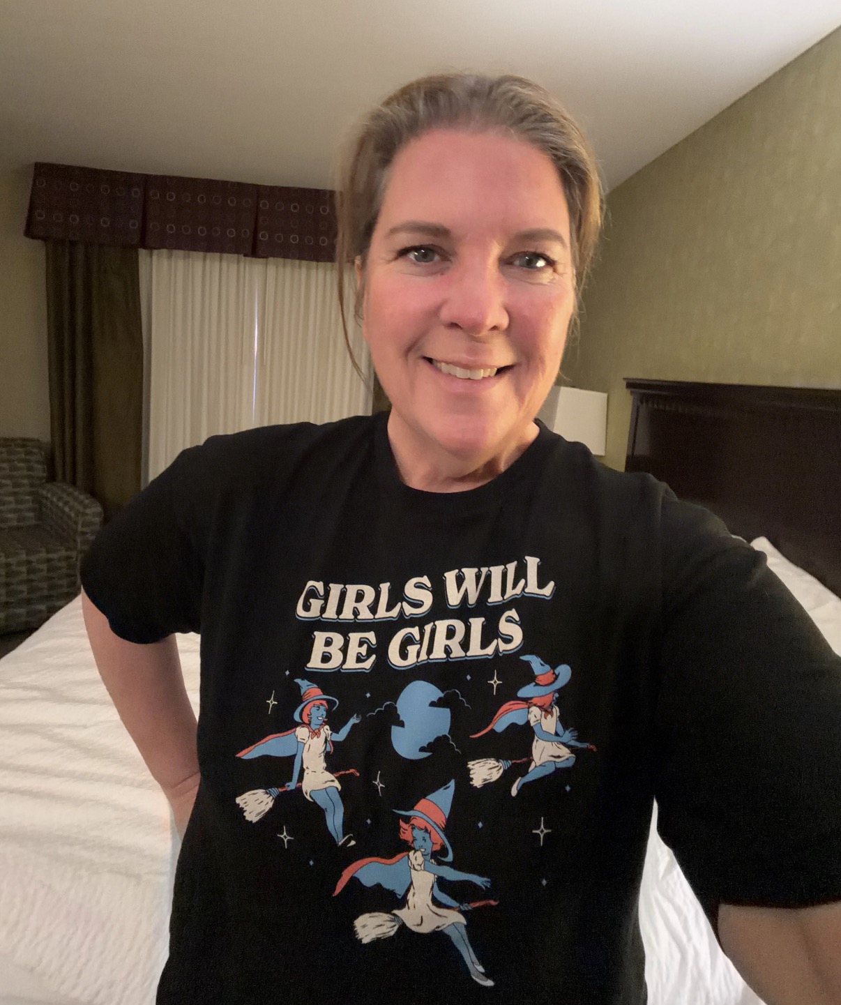 Therese Marzouk on location in a hotel wearing a black t-shirt that says "girls will be girls" with three flying witches.